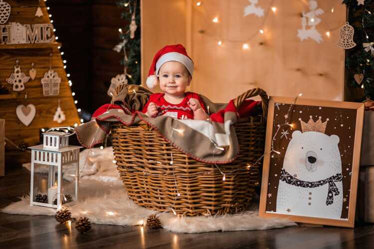 Adorable baby in Santa hat and sweater sitting in basket in decorated room with garlands